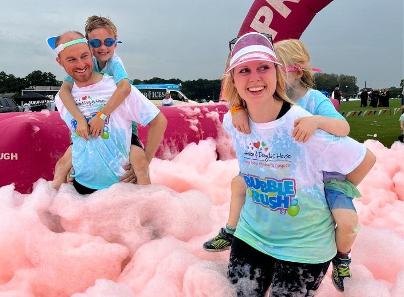 Two smiling adults carrying children on their backs as they walk through pink foamy bubbles.