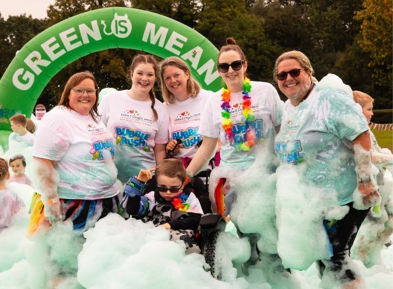 Five smiling women and a boy in a wheelchair surrounded by green bubble foam.
